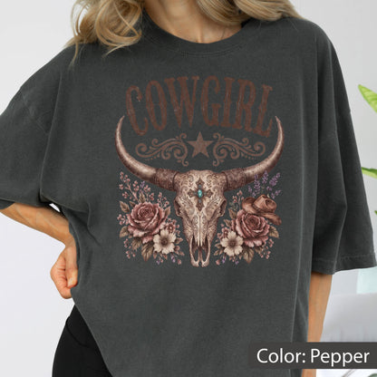 May include: A charcoal gray t-shirt featuring a graphic print of a steer skull with long horns, centered below the text COWGIRL. The design includes a star, floral accents with roses, and a small cowboy hat, all rendered in muted earth tones.
