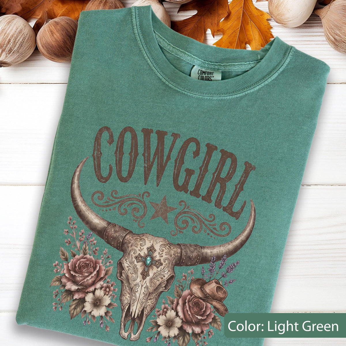 May include: A light green short-sleeve t-shirt with a crew neckline. The graphic features the word COWGIRL in brown letters above a steer skull with a cross, floral accents, a star, and a small hat. The Comfort Colors brand tag is visible.
