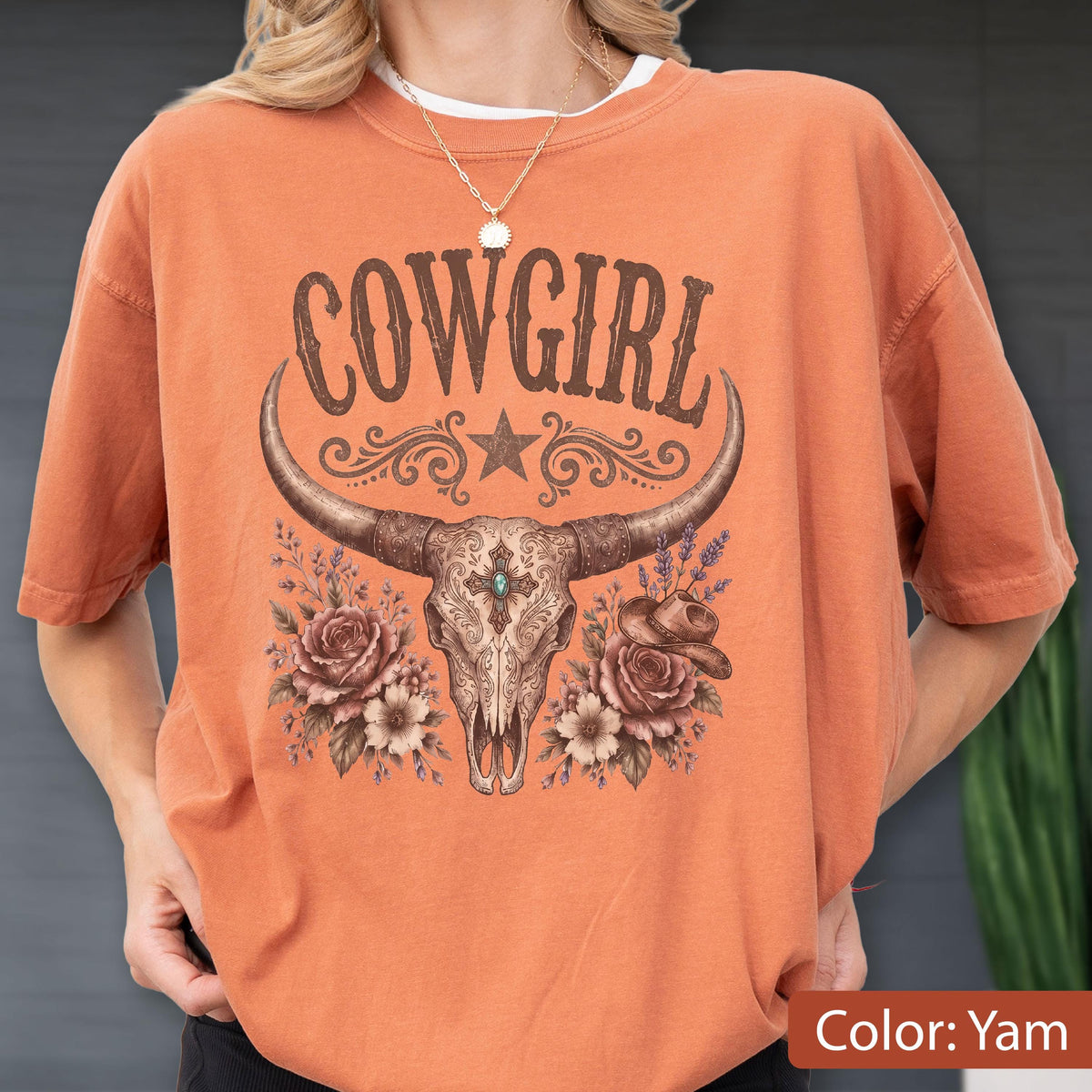 May include: A yam-colored t-shirt featuring a graphic print of a longhorn skull centered above the text COWGIRL. The design includes decorative scrollwork, a star, roses, and a small hat. A silver-toned chain necklace rests at the neckline.