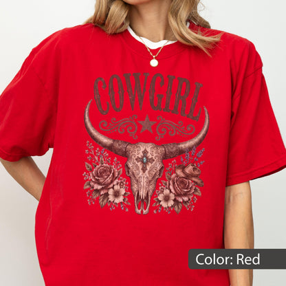 May include: A bright red short-sleeve t-shirt featuring a graphic print of a longhorn skull adorned with roses and a small cowboy hat. The text COWGIRL is printed in a distressed brown font above the skull, which includes a central cross detail.