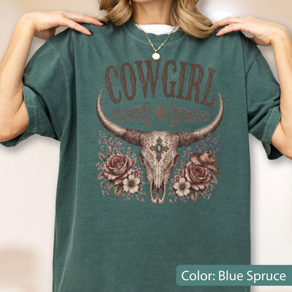 May include: A blue spruce short-sleeve t-shirt featuring a graphic print of a longhorn skull, roses, and a cowboy hat. The text COWGIRL is printed in a brown western-style font above the skull, which is decorated with ornate filigree patterns.