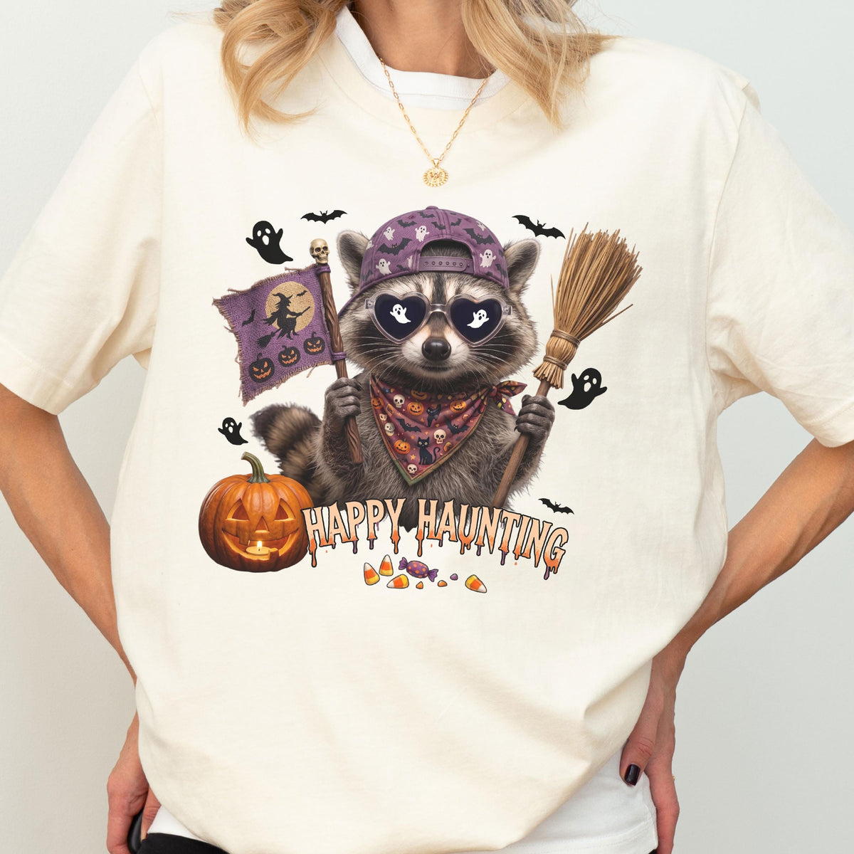 May include: A cream-colored t-shirt featuring a graphic of a raccoon wearing a purple baseball cap, heart-shaped sunglasses with ghost reflections, and a patterned bandana. The raccoon holds a flag with a witch and a broomstick near a pumpkin.