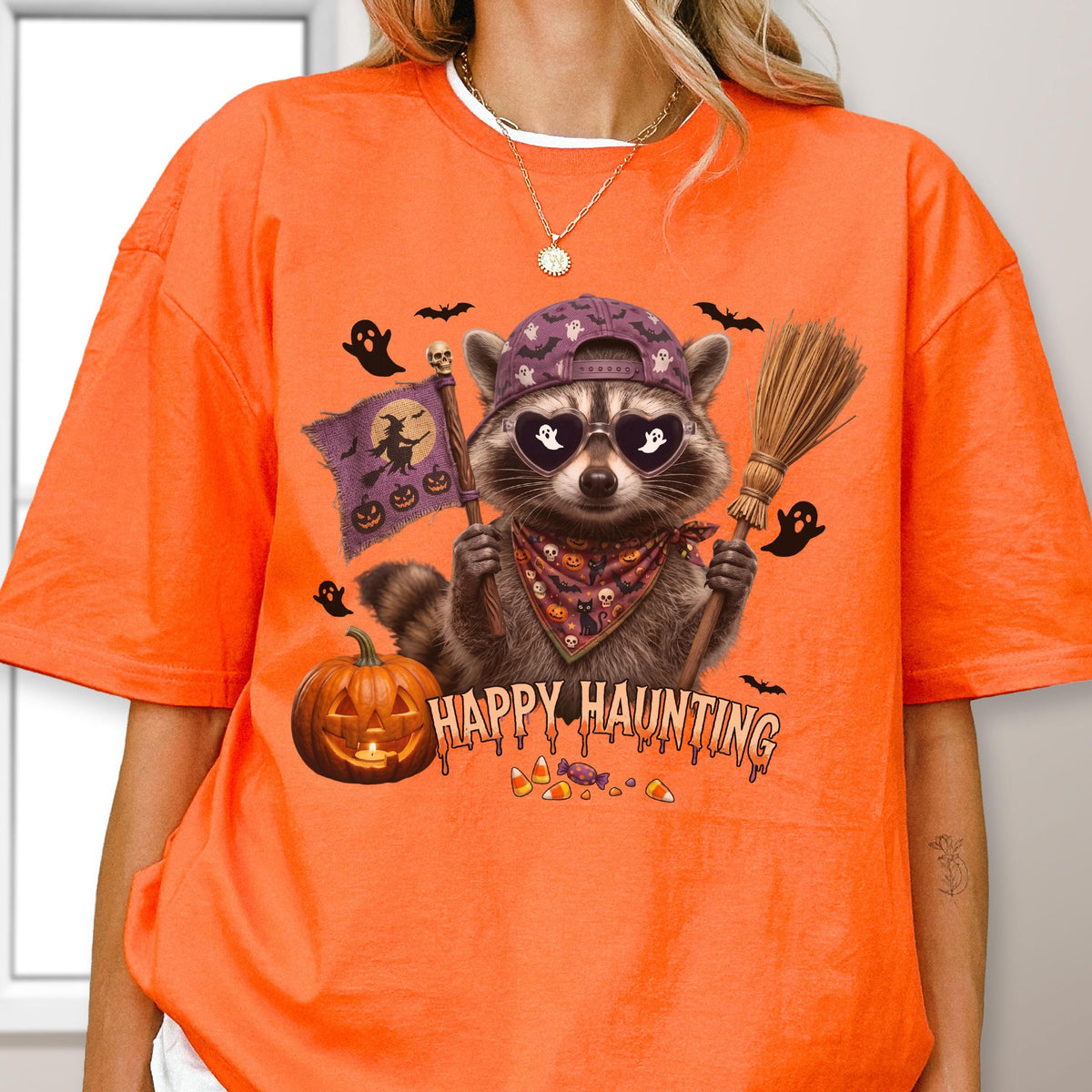 May include: An orange t-shirt featuring a graphic of a raccoon wearing a purple baseball cap, heart-shaped sunglasses with ghost reflections, and a patterned bandana. The raccoon holds a flag and a broom near a pumpkin with the text HAPPY HAUNTING.