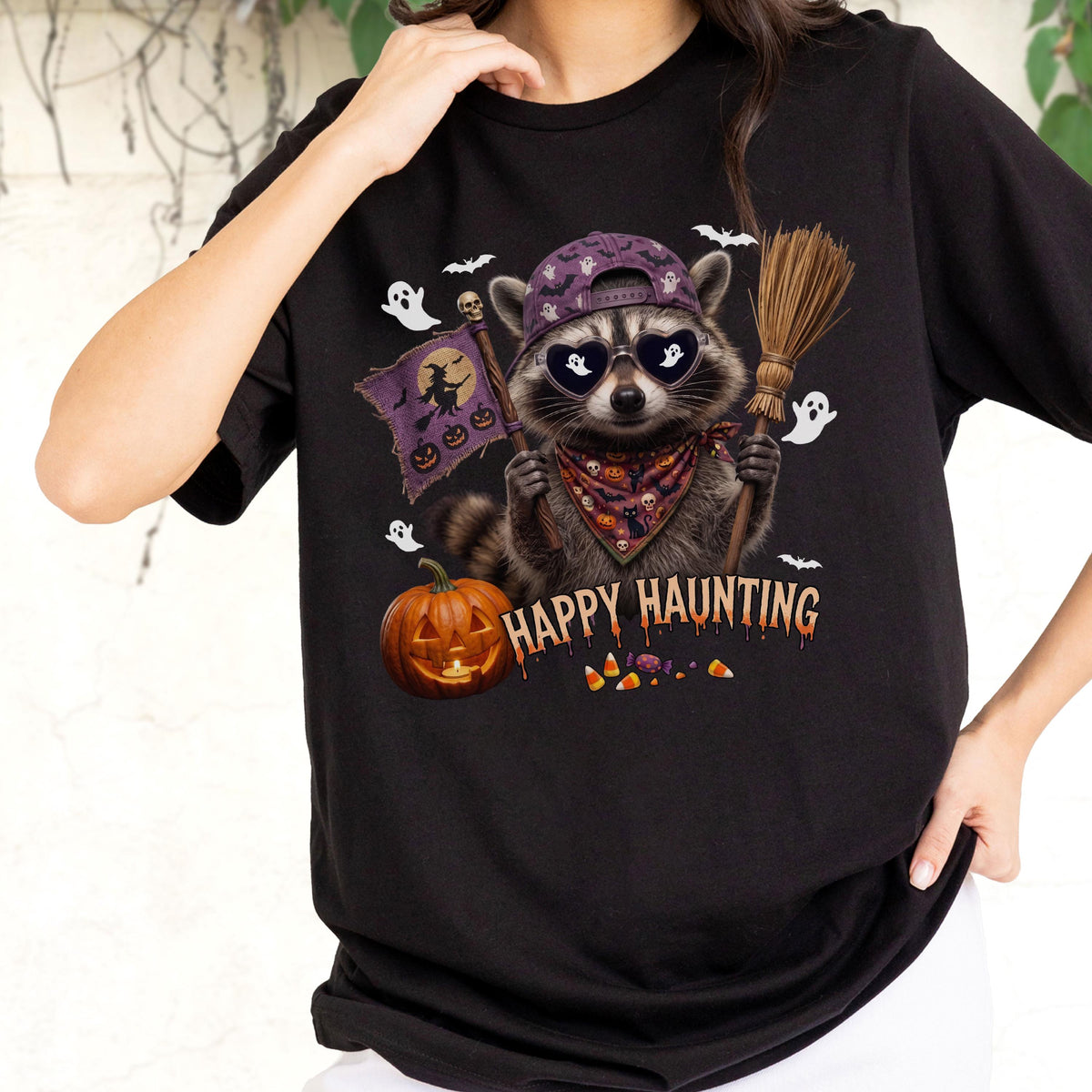 May include: A black t-shirt featuring a graphic of a raccoon wearing a purple baseball cap, heart-shaped sunglasses, and a patterned bandana. The raccoon holds a broom and a flag with a witch silhouette. Text reads HAPPY HAUNTING.