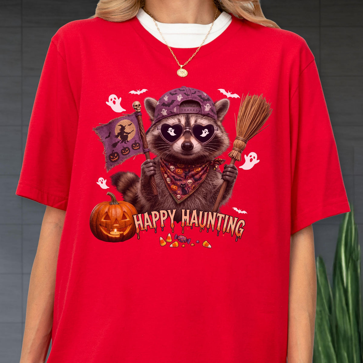 May include: A bright red t-shirt featuring a graphic of a raccoon wearing a purple baseball cap and heart-shaped sunglasses. The raccoon holds a broom and a flag with a witch silhouette. Text reads Happy Haunting above a carved orange pumpkin.