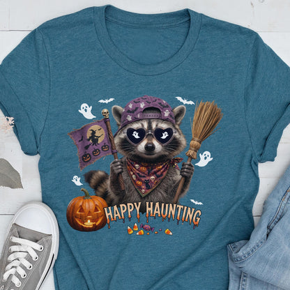 May include: A teal t-shirt featuring a graphic of a raccoon wearing a purple bat-patterned baseball cap and heart-shaped sunglasses with ghost reflections. The raccoon holds a broom and a flag with a witch silhouette, next to a pumpkin and the text HAPPY HAUNTING.
