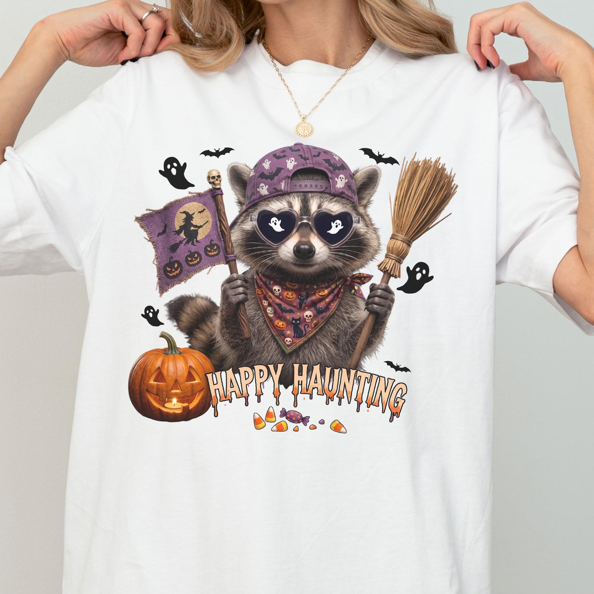 May include: A white t-shirt featuring a graphic of a raccoon wearing a purple baseball cap, heart-shaped sunglasses, and a patterned bandana. The raccoon holds a broom and a flag with a witch silhouette. Text reads HAPPY HAUNTING with a pumpkin.