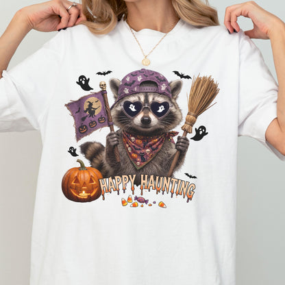 May include: A white t-shirt featuring a graphic of a raccoon wearing a purple baseball cap, heart-shaped sunglasses, and a patterned bandana. The raccoon holds a broom and a flag with a witch silhouette. Text reads HAPPY HAUNTING with a pumpkin.