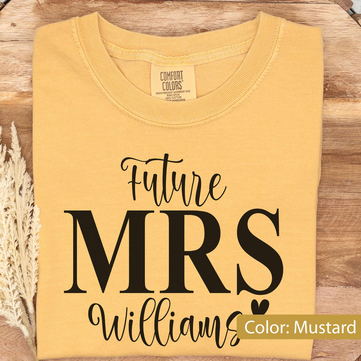 May include: A mustard yellow t-shirt featuring the black printed text Future MRS Williams with a small heart icon. The shirt has a crew neckline and a Comfort Colors brand label on the inside collar. The design is centered on the front of the garment.