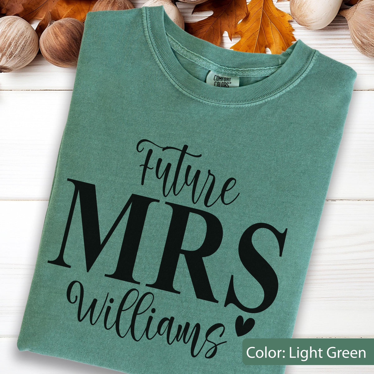 May include: A light green crew neck t-shirt featuring the black text Future MRS Williams with a small heart icon. The shirt has a Comfort Colors brand label visible at the inner neckline and is made of a soft, garment-dyed cotton fabric.
