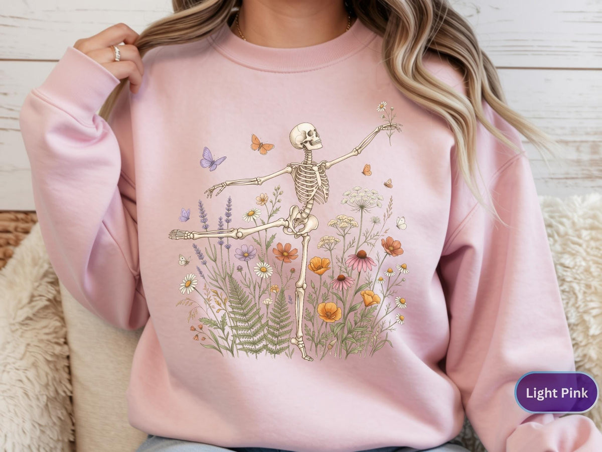 May include: A light pink crewneck sweatshirt featuring a graphic print of a skeleton dancing amidst a field of colorful wildflowers, ferns, and small butterflies. The skeleton is positioned in a ballet-like pose, reaching out with one arm.
