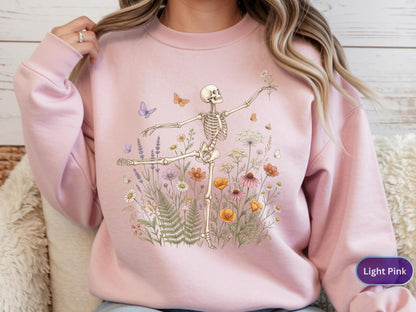 May include: A light pink crewneck sweatshirt featuring a graphic print of a skeleton dancing amidst a field of colorful wildflowers, ferns, and small butterflies. The skeleton is positioned in a ballet-like pose, reaching out with one arm.