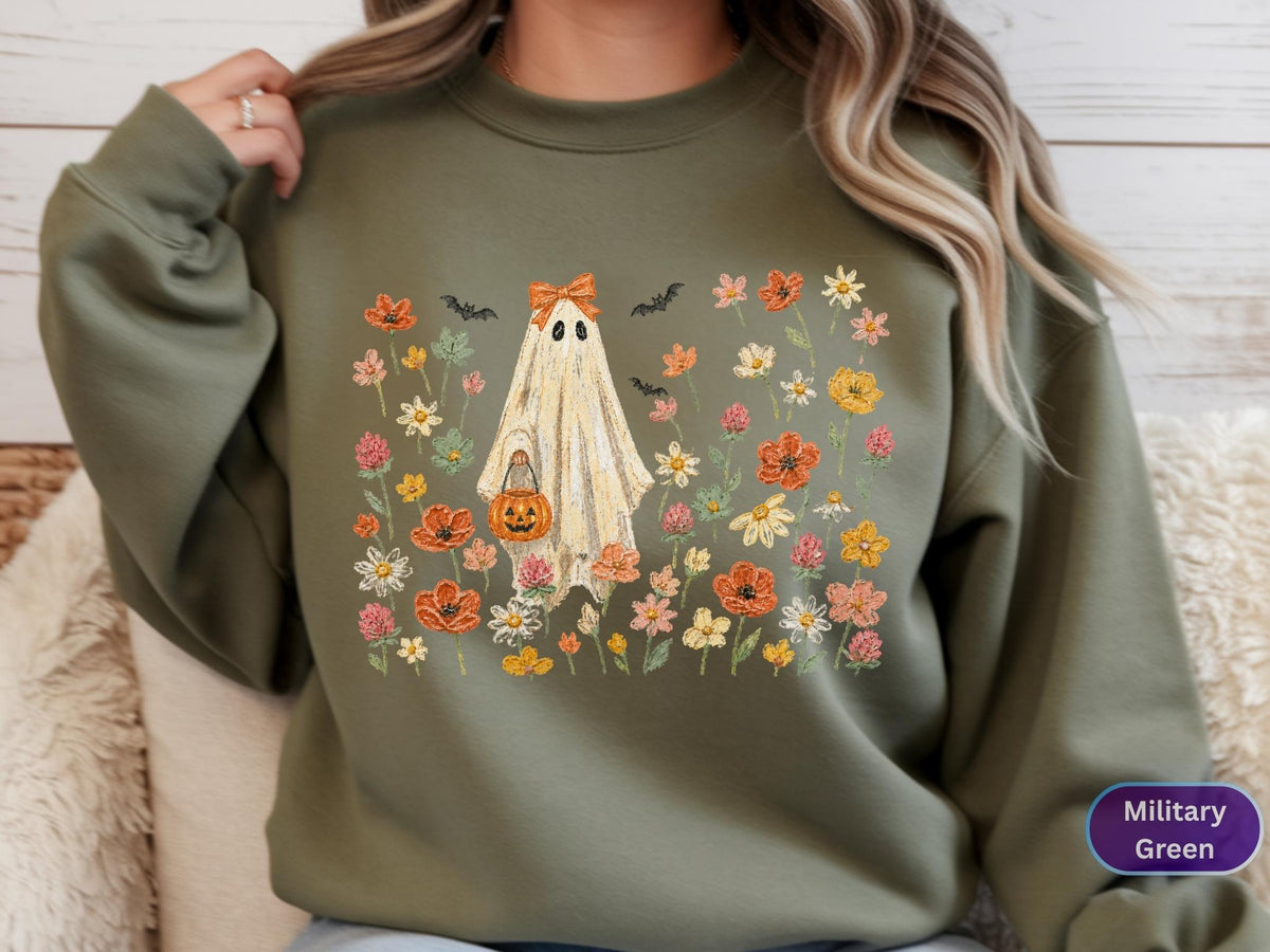 May include: A military green crewneck sweatshirt featuring a central graphic of a ghost wearing an orange bow and holding a jack-o'-lantern bucket. The ghost is surrounded by a colorful field of various wildflowers and small black bats.