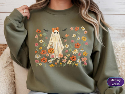 May include: A military green crewneck sweatshirt featuring a central graphic of a ghost wearing an orange bow and holding a jack-o'-lantern bucket. The ghost is surrounded by a colorful field of various wildflowers and small black bats.
