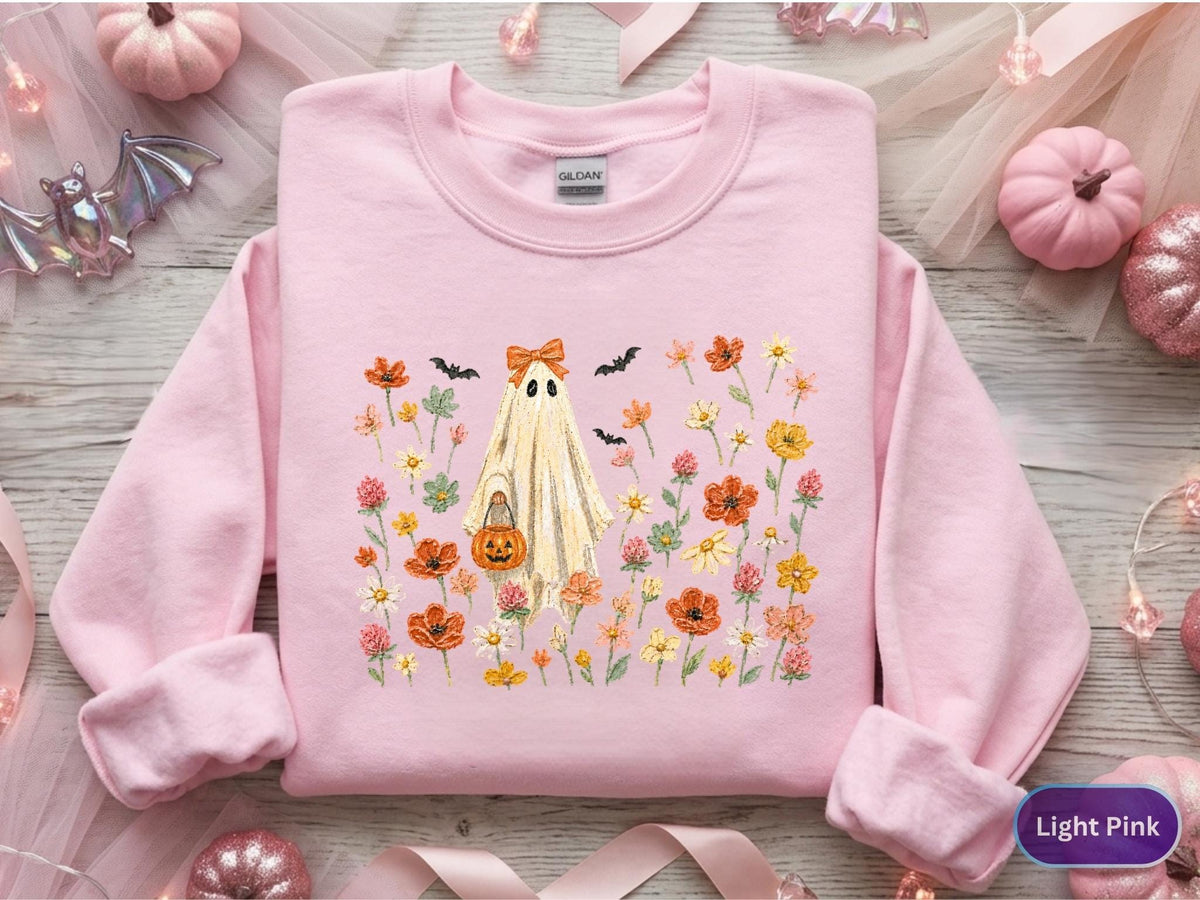 May include: A light pink crewneck sweatshirt featuring a central graphic of a ghost wearing an orange bow and holding a jack-o'-lantern bucket. The ghost is surrounded by a colorful field of orange, yellow, and pink wildflowers and small black bats.