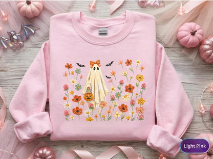 May include: A light pink crewneck sweatshirt featuring a central graphic of a ghost wearing an orange bow and holding a jack-o'-lantern bucket. The ghost is surrounded by a colorful field of orange, yellow, and pink wildflowers and small black bats.