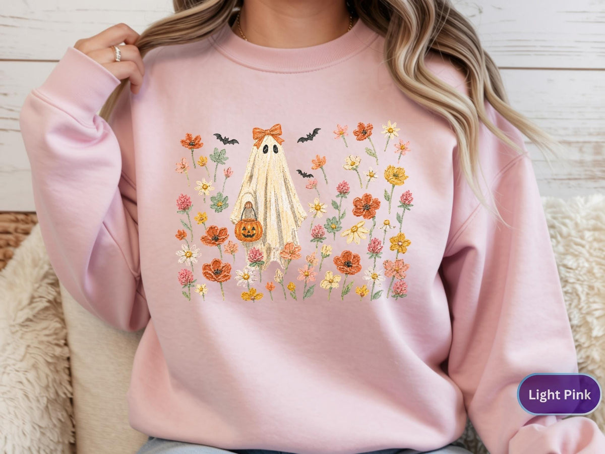 May include: A light pink crewneck sweatshirt featuring a central graphic of a ghost wearing an orange bow, holding a jack-o'-lantern bucket, surrounded by a colorful field of wildflowers and small black bats in shades of orange, yellow, and green.