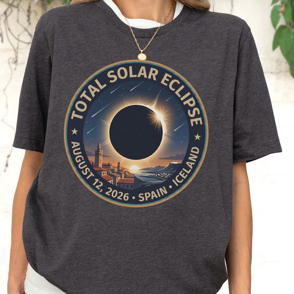 May include: A charcoal gray t-shirt featuring a circular graphic of a total solar eclipse. The text reads TOTAL SOLAR ECLIPSE AUGUST 12, 2026 SPAIN ICELAND. The design depicts a dark moon blocking the sun over a coastal town with buildings.