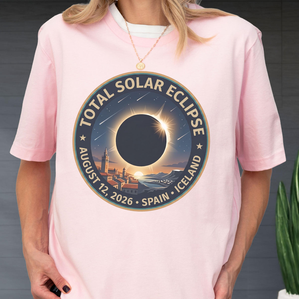 May include: A light pink short-sleeve t-shirt featuring a circular graphic of a total solar eclipse over a coastal town. The text reads TOTAL SOLAR ECLIPSE AUGUST 12, 2026 SPAIN ICELAND. The design includes a dark sun, stars, and a cityscape.