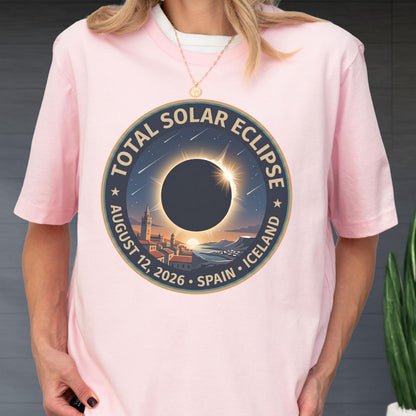May include: A light pink short-sleeve t-shirt featuring a circular graphic of a total solar eclipse over a coastal town. The text reads TOTAL SOLAR ECLIPSE AUGUST 12, 2026 SPAIN ICELAND. The design includes a dark sun, stars, and a cityscape.