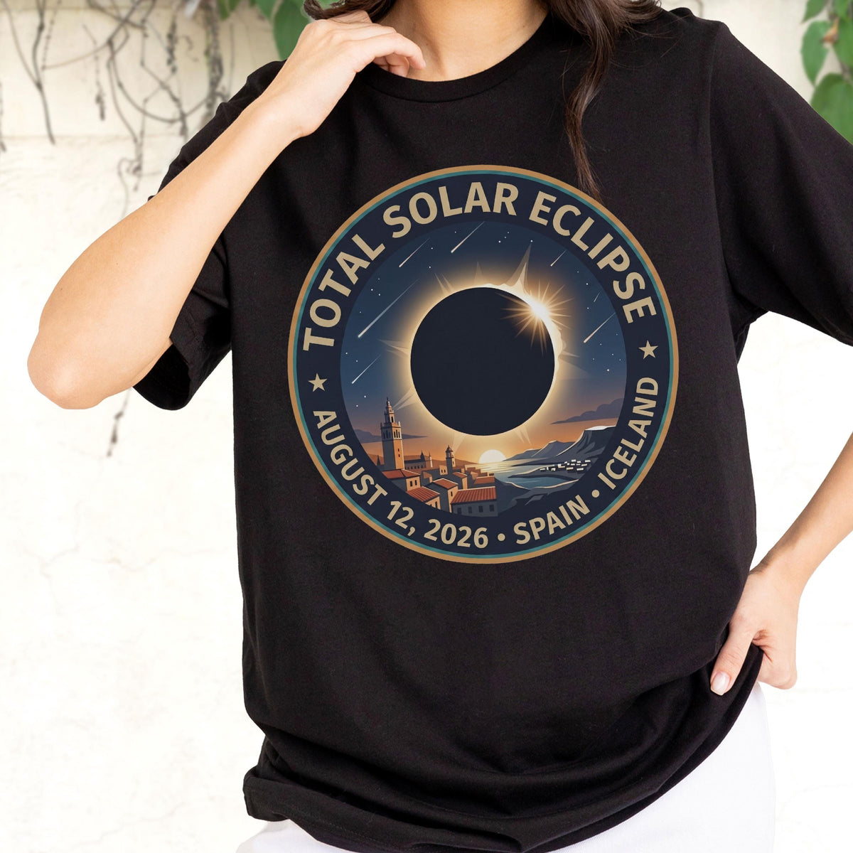 May include: A black t-shirt featuring a circular graphic of a total solar eclipse over a coastal town. The text reads TOTAL SOLAR ECLIPSE, AUGUST 12, 2026, SPAIN, ICELAND. The design includes a dark sun, a bright corona, and falling stars.