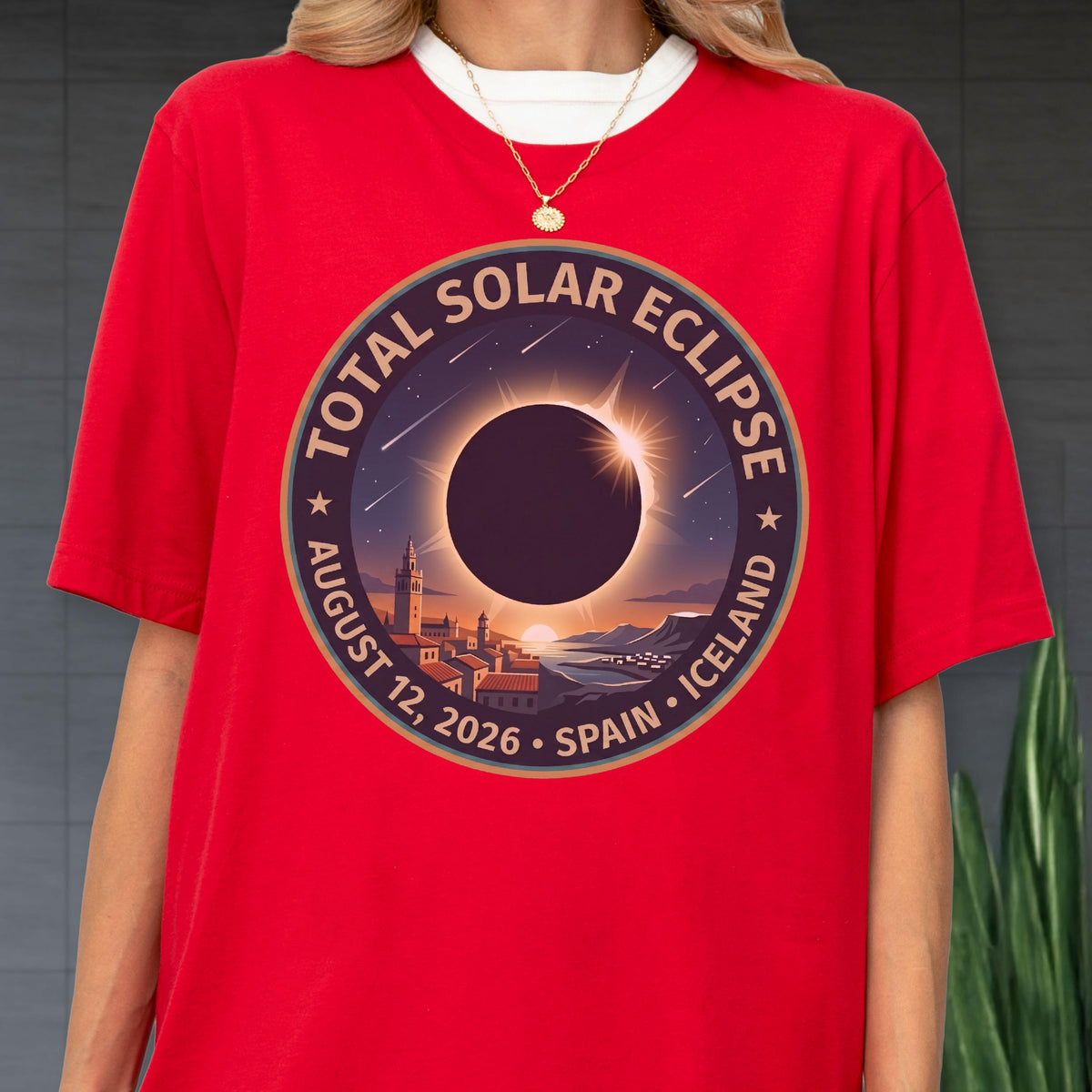 May include: A bright red t-shirt featuring a circular graphic of a total solar eclipse over a coastal town. The text reads Total Solar Eclipse, August 12, 2026, Spain, Iceland. The design includes a dark sun, a bright corona, and stars.