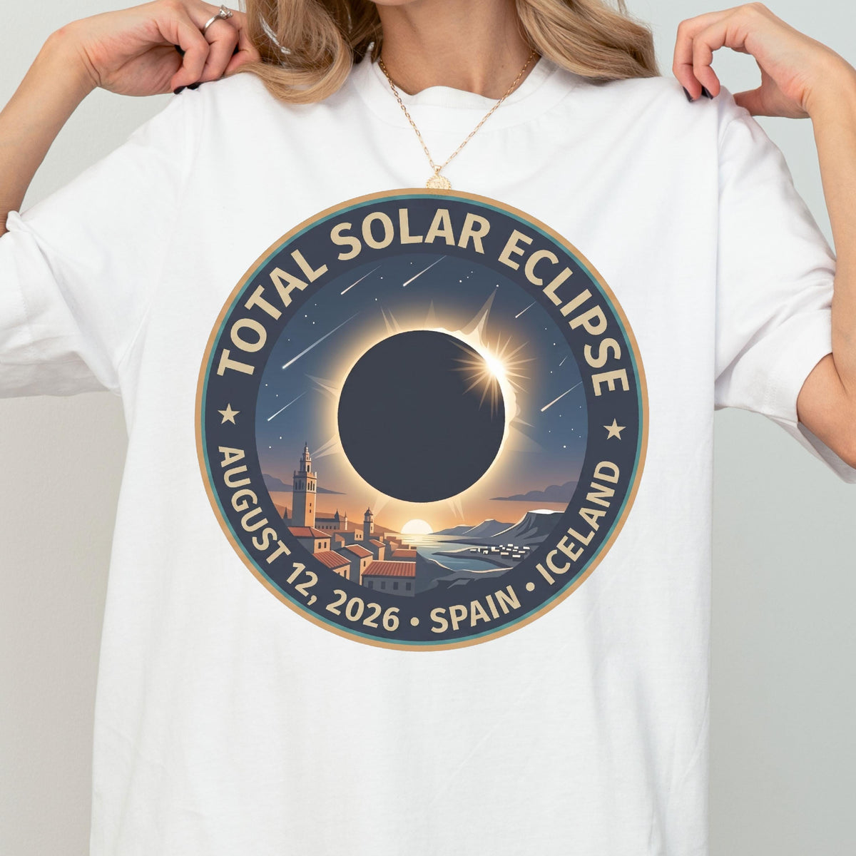 May include: A white t-shirt featuring a circular graphic of a total solar eclipse. The design includes the text TOTAL SOLAR ECLIPSE AUGUST 12, 2026 SPAIN ICELAND, a dark moon silhouette, a glowing sun corona, shooting stars, and a coastal town landscape.
