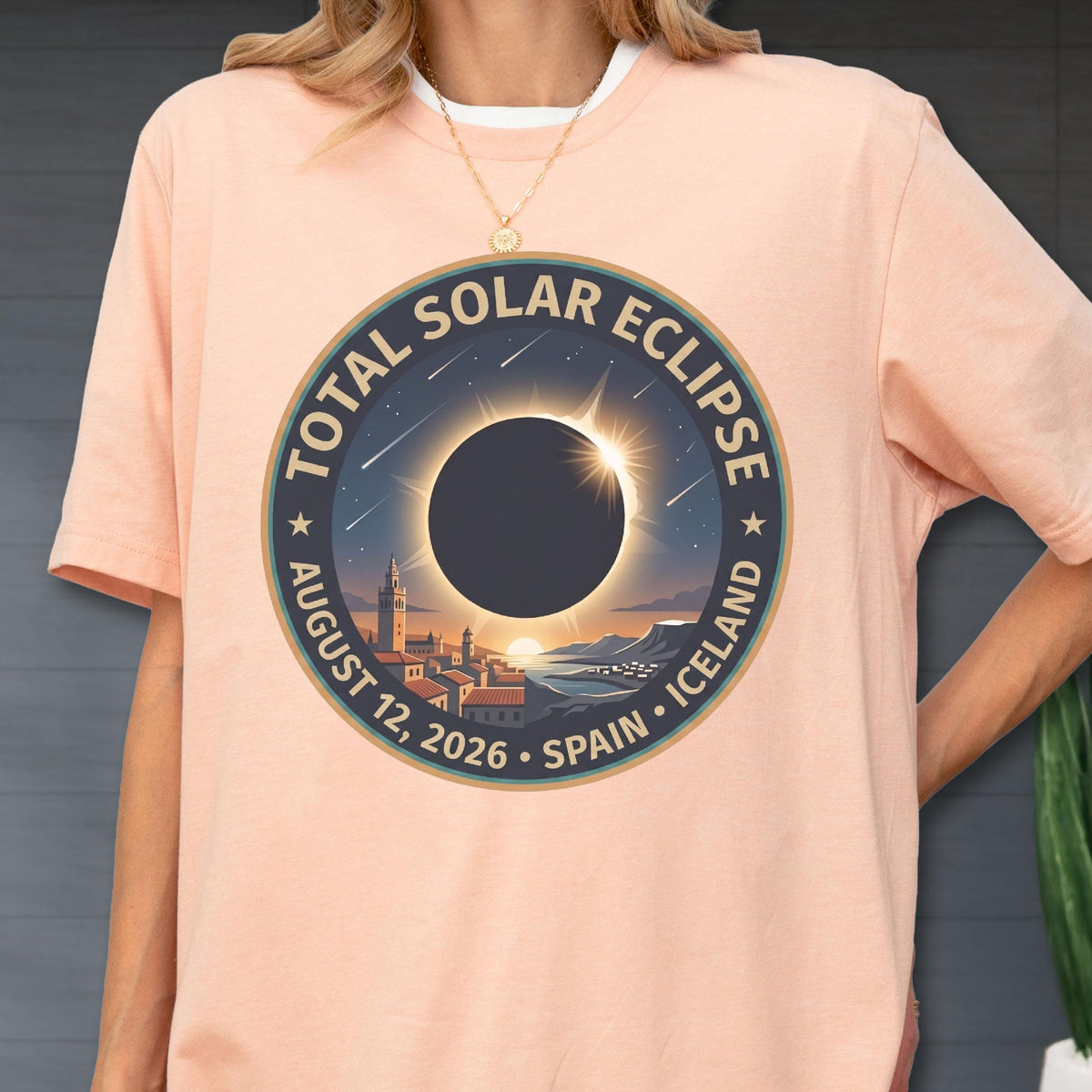 May include: A peach-colored t-shirt featuring a circular graphic of a total solar eclipse over a coastal town. The text reads TOTAL SOLAR ECLIPSE AUGUST 12, 2026 SPAIN ICELAND. The design includes a dark moon, a bright corona, and shooting stars.