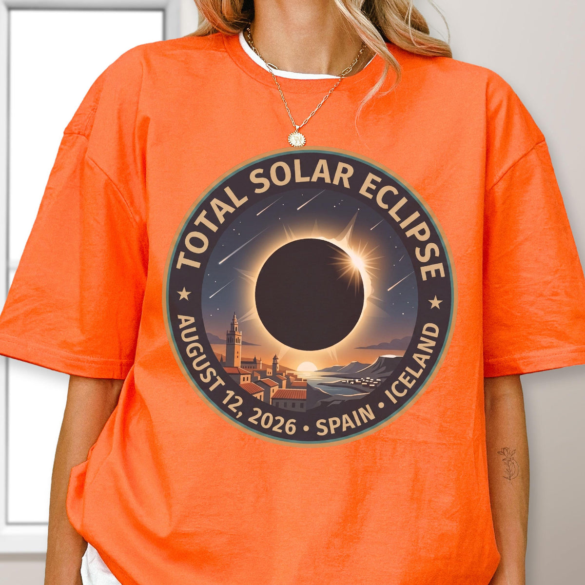 May include: A bright orange short-sleeve t-shirt featuring a circular graphic of a total solar eclipse over a coastal town. The text reads TOTAL SOLAR ECLIPSE AUGUST 12, 2026 SPAIN ICELAND. The design includes a dark sun, light rays, and stars.