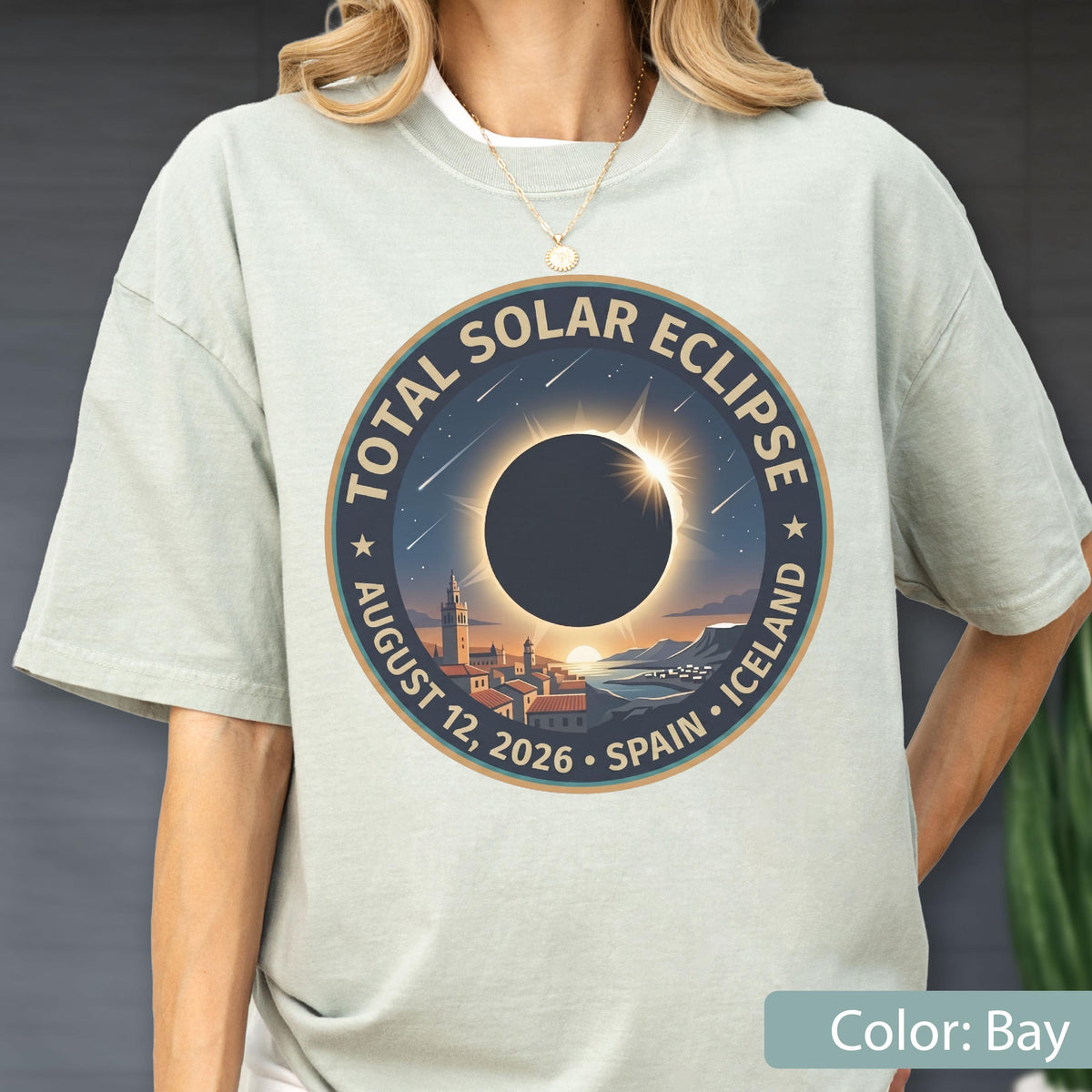 May include: A light bay-colored t-shirt featuring a circular graphic of a total solar eclipse. The design includes the text "TOTAL SOLAR ECLIPSE" and "AUGUST 12, 2026 • SPAIN • ICELAND" surrounding an illustration of a dark sun over a coastal town.
