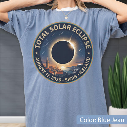 May include: A blue jean colored t-shirt featuring a circular graphic of a total solar eclipse over a coastal town. The text reads TOTAL SOLAR ECLIPSE AUGUST 12, 2026 SPAIN ICELAND. The design includes a dark sun silhouette, rays, and stars.