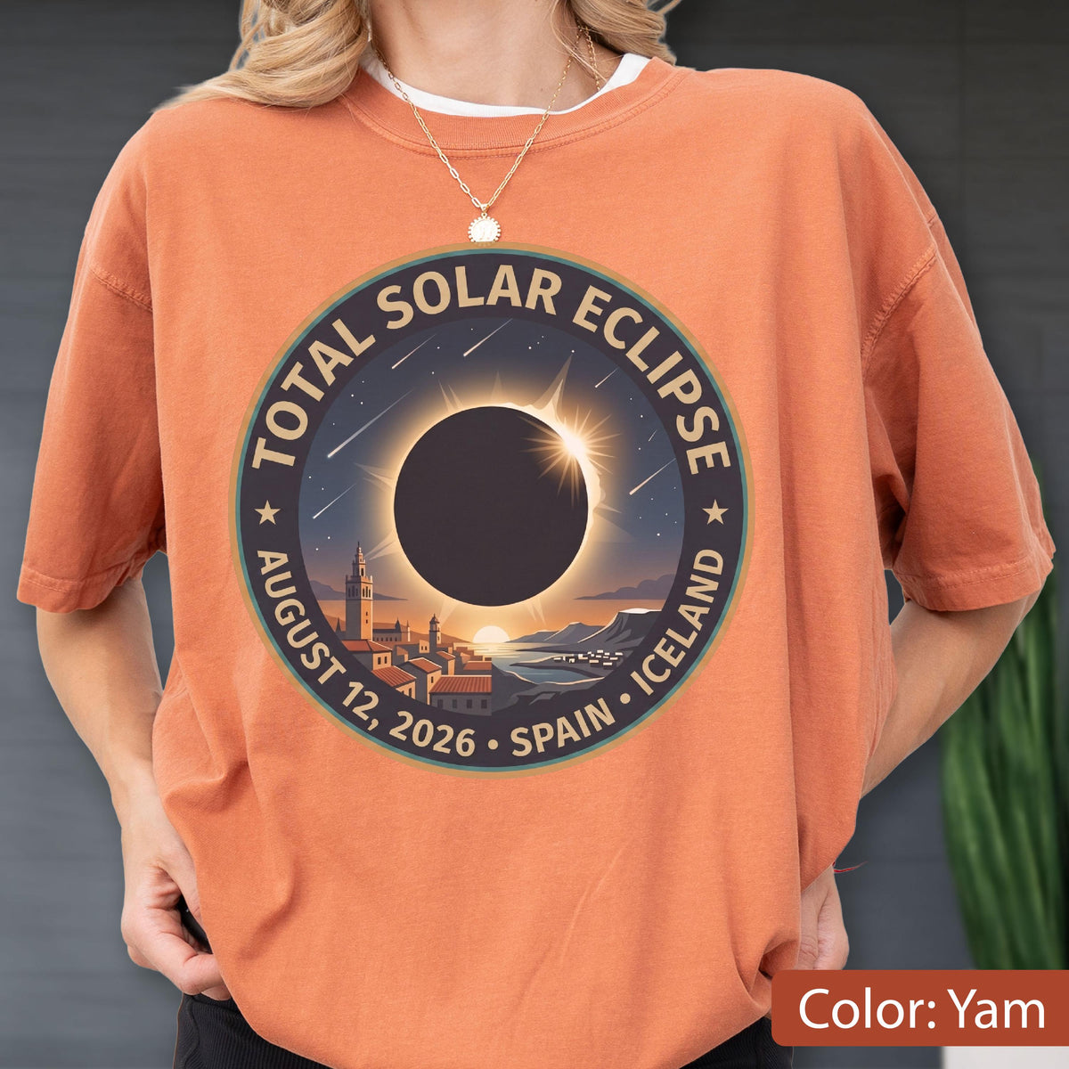 May include: A yam-colored t-shirt featuring a circular graphic of a total solar eclipse over a coastal town. The text reads TOTAL SOLAR ECLIPSE AUGUST 12, 2026 SPAIN ICELAND. The design includes a dark sun, a bright corona, and stars.