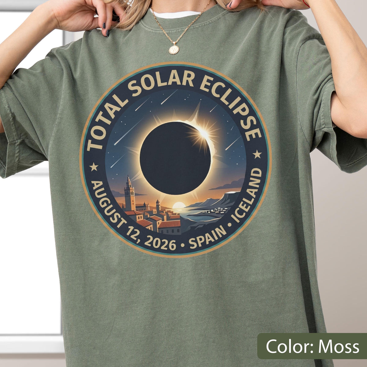 May include: A moss green t-shirt featuring a circular graphic of a total solar eclipse over a coastal town. The text reads TOTAL SOLAR ECLIPSE, AUGUST 12, 2026, SPAIN, ICELAND. The design includes a dark sun, bright corona, and falling stars.