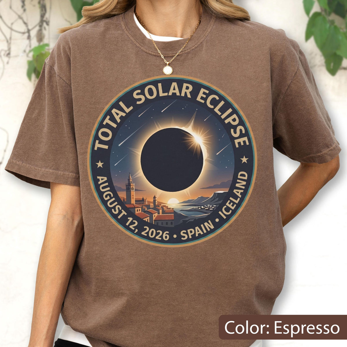 May include: An espresso-colored short-sleeve t-shirt featuring a circular graphic of a total solar eclipse over a coastal town. The text reads Total Solar Eclipse, August 12, 2026, Spain, Iceland. The design includes a dark sun, rays, and stars.