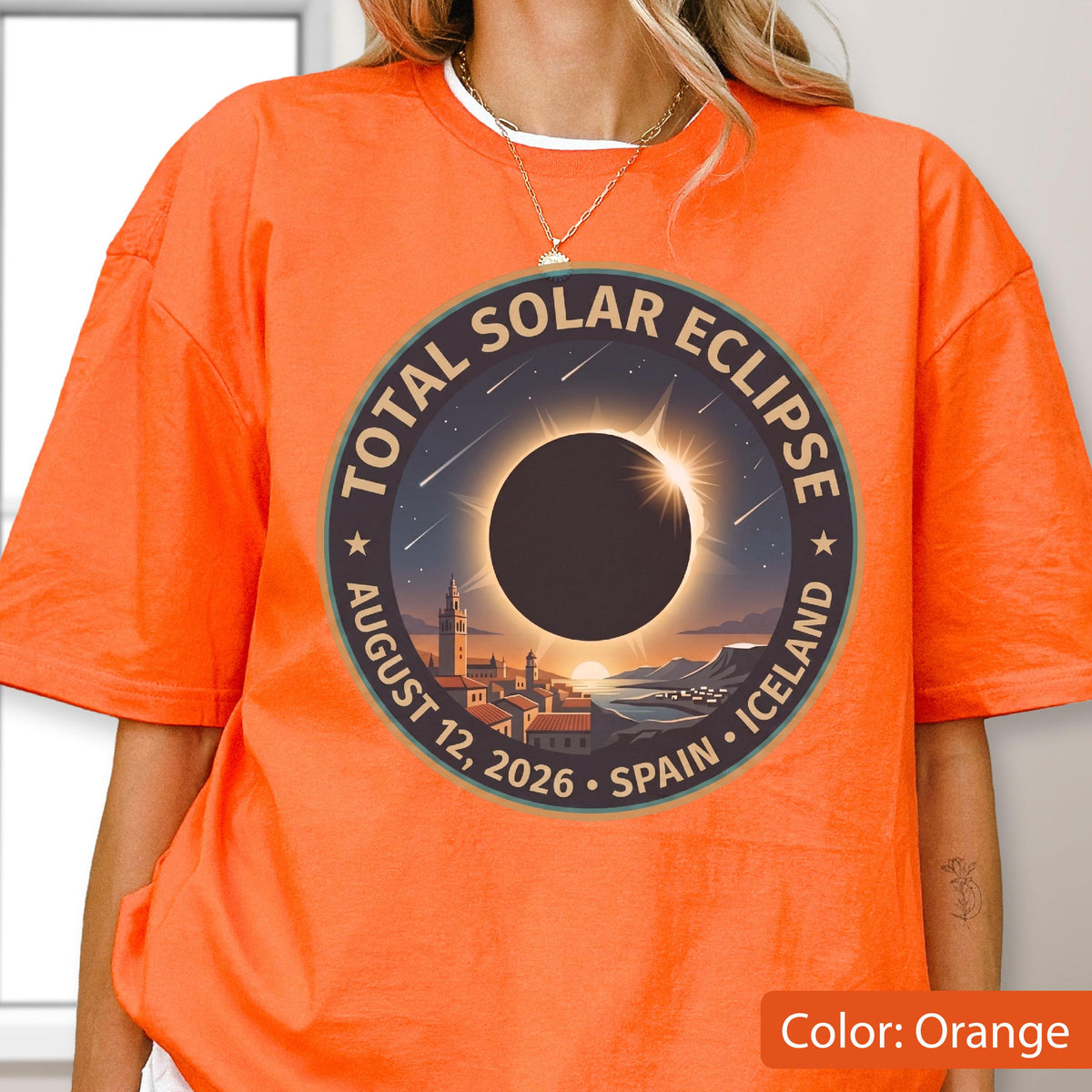 May include: A bright orange t-shirt featuring a circular graphic of a total solar eclipse over a coastal town. The text reads Total Solar Eclipse, August 12, 2026, Spain, Iceland. The design includes a dark sun silhouette, stars, and buildings.