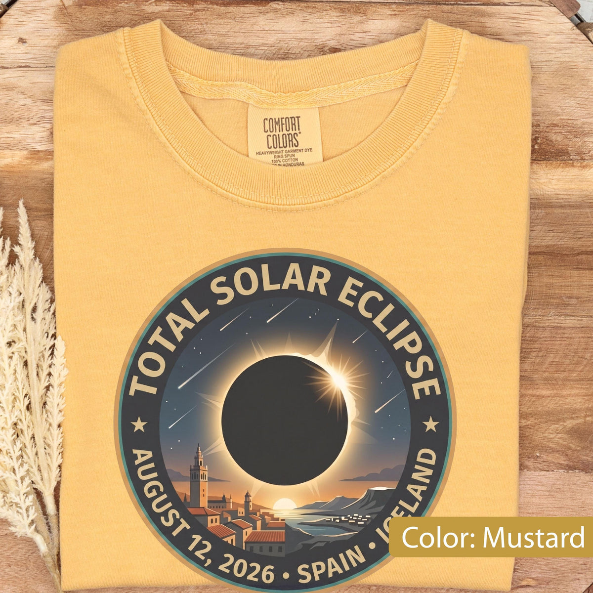May include: A mustard yellow t-shirt featuring a circular graphic of a total solar eclipse over a coastal town. The text reads TOTAL SOLAR ECLIPSE AUGUST 12, 2026 SPAIN ICELAND. The design includes a dark moon silhouette, sun rays, and stars.