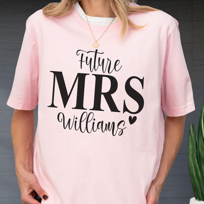 May include: A light pink short-sleeved t-shirt featuring black text that reads Future MRS Williams with a small heart icon. The shirt has a crew neckline and is made of a solid-colored fabric, providing a simple and clean look for casual wear.