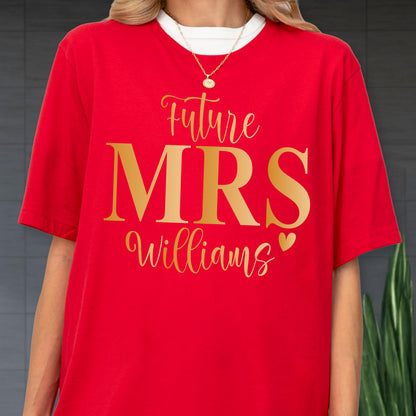 May include: A bright red short-sleeved t-shirt featuring the gold text Future MRS Williams with a small gold heart icon. The shirt is worn over a white crew neck base layer and is paired with a thin gold chain necklace with a circular pendant.