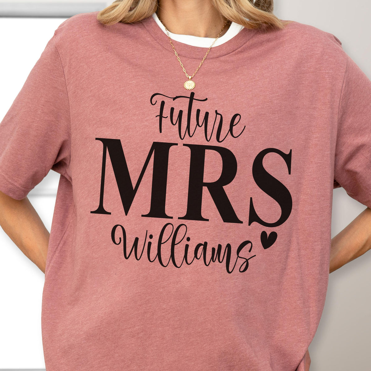 May include: A dusty rose short-sleeved t-shirt featuring black text that reads Future MRS Williams with a small heart icon. The shirt has a crew neckline and is worn with a gold chain necklace featuring a small circular pendant.