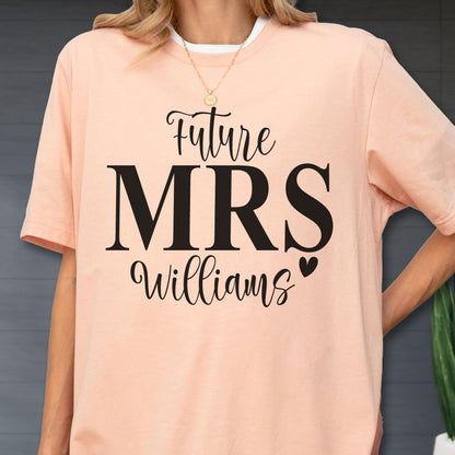 May include: A peach-colored short-sleeve t-shirt featuring black text that reads Future MRS Williams with a small heart icon. The shirt has a crew neckline and a relaxed fit, showcasing a clear graphic print centered on the front of the garment.