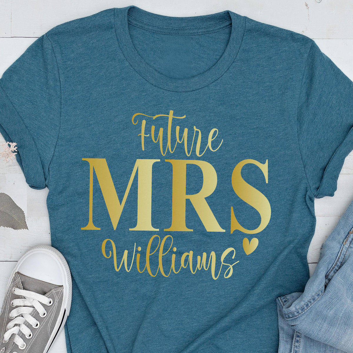 May include: A teal short-sleeved t-shirt featuring gold text that reads Future MRS Williams with a small heart icon. The shirt has a crew neckline and is displayed alongside a gray sneaker and a portion of blue denim fabric.