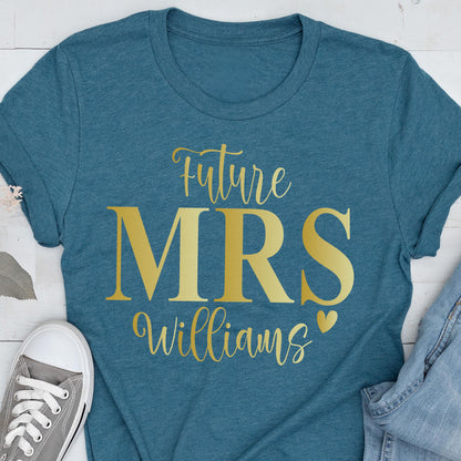 May include: A teal short-sleeved t-shirt featuring gold text that reads Future MRS Williams with a small heart icon. The shirt has a crew neckline and is displayed alongside a gray sneaker and a portion of blue denim fabric.
