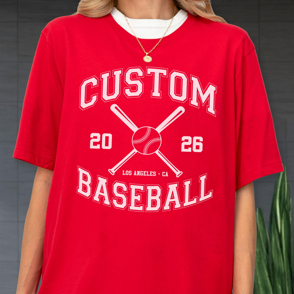 May include: A bright red short-sleeved t-shirt featuring a white graphic print. The design includes the text CUSTOM BASEBALL, the numbers 20 and 26, two crossed baseball bats, and a baseball. The text LOS ANGELES - CA is printed below the ball.