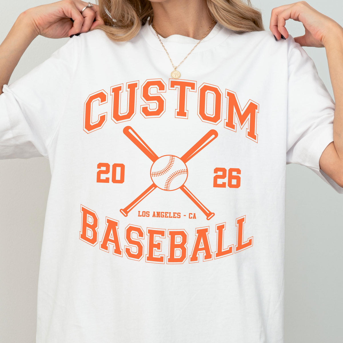 May include: A white t-shirt featuring an orange graphic print with the text CUSTOM BASEBALL, the numbers 20 and 26, and the location LOS ANGELES - CA. The design includes a central illustration of a baseball crossed by two baseball bats.