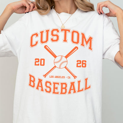 May include: A white t-shirt featuring an orange graphic print with the text CUSTOM BASEBALL, the numbers 20 and 26, and the location LOS ANGELES - CA. The design includes a central illustration of a baseball crossed by two baseball bats.