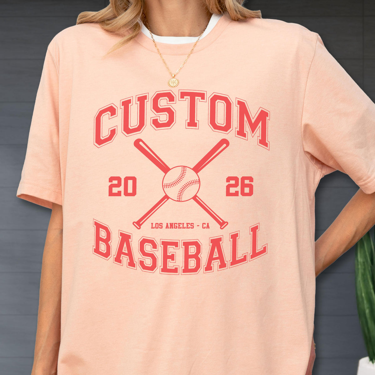 May include: A peach-colored short-sleeve t-shirt featuring a red graphic print. The design includes the text CUSTOM BASEBALL, the numbers 20 and 26, the location LOS ANGELES - CA, and an illustration of a baseball centered between two crossed bats.