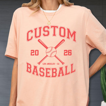 May include: A peach-colored short-sleeve t-shirt featuring a red graphic print. The design includes the text CUSTOM BASEBALL, the numbers 20 and 26, the location LOS ANGELES - CA, and an illustration of a baseball centered between two crossed bats.