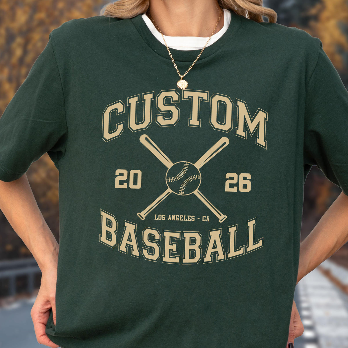 May include: A dark green short-sleeve t-shirt featuring a tan graphic print with the text CUSTOM BASEBALL, the numbers 20 and 26, and the location LOS ANGELES - CA, centered around an illustration of two crossed baseball bats and a baseball.