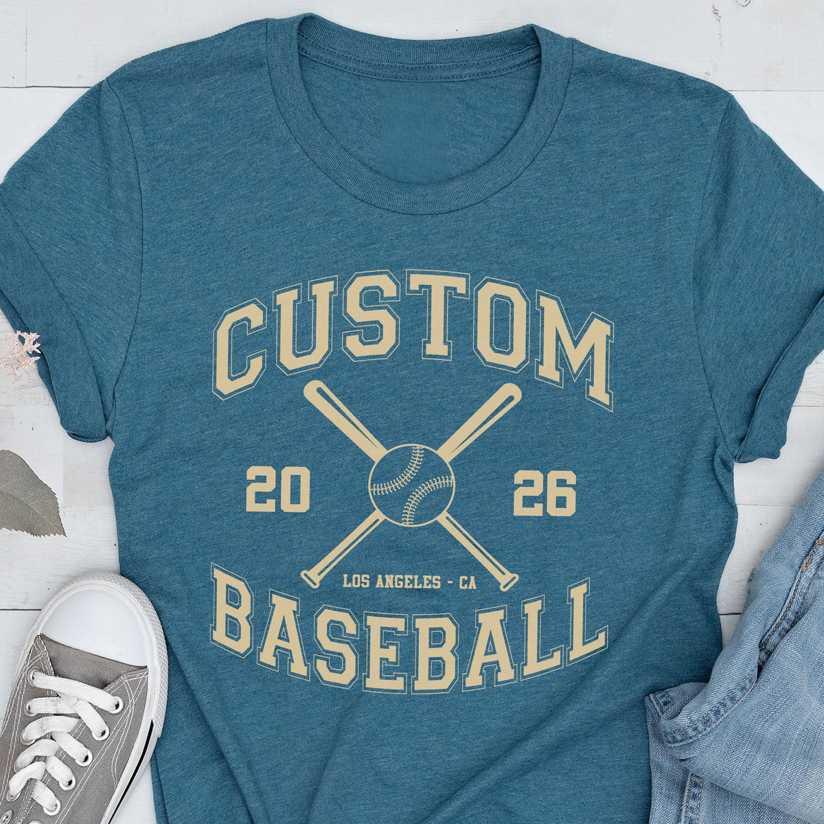May include: A teal short-sleeve t-shirt featuring a cream-colored graphic with the text CUSTOM BASEBALL, the numbers 20 and 26, and an illustration of two crossed baseball bats behind a baseball, with the text LOS ANGELES - CA below the ball.