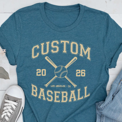 May include: A teal short-sleeve t-shirt featuring a cream-colored graphic with the text CUSTOM BASEBALL, the numbers 20 and 26, and an illustration of two crossed baseball bats behind a baseball, with the text LOS ANGELES - CA below the ball.